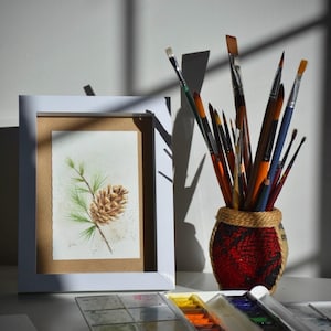 May include: A white picture frame containing a watercolour painting of a brown pinecone on a green pine branch. Next to the frame is a textured red and black woven cup filled with various paintbrushes and a plastic watercolour paint palette.