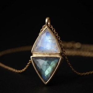 May include: A gold pendant necklace featuring two stacked triangular faceted gemstones. The top stone is a translucent white moonstone with blue iridescence, and the bottom stone is a dark grey labradorite with green and blue flashes.