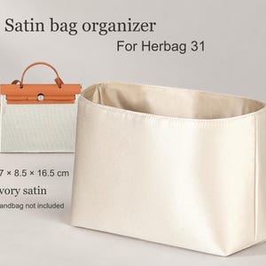May include: An ivory satin bag organizer for Herbag 31, measuring 27 x 8.5 x 16.5 cm. The rectangular insert features a smooth, lustrous finish and is shown alongside a small tan and white handbag for scale and reference.