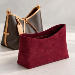 May include: A deep red suede pouch with a rectangular base and an open top sits in front of a dark brown leather tote bag. The tote features light tan leather trim, gold-tone hardware, and a matching leather luggage tag attached to the handle.