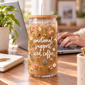 May include: A clear glass tumbler filled with iced coffee, featuring a bamboo lid, a glass straw, and a floral pattern with the white cursive text "emotional support iced coffee" printed on the front.