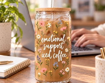 May include: A clear glass tumbler filled with iced coffee, featuring a bamboo lid, a glass straw, and a floral pattern with the white cursive text "emotional support iced coffee" printed on the front.