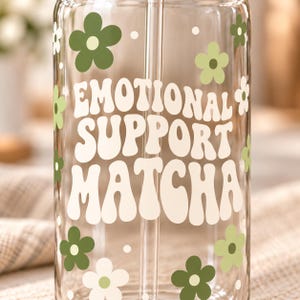 May include: A clear glass tumbler with a straw featuring the text EMOTIONAL SUPPORT MATCHA in large cream-coloured letters, surrounded by various small flowers in shades of dark green, light green, and cream, along with small white dots.