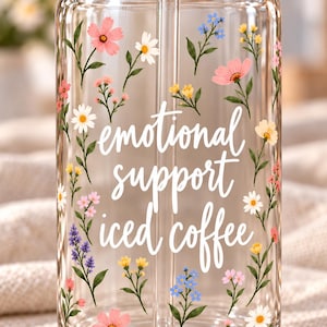 May include: A clear glass tumbler with a straw featuring a floral pattern of pink, yellow, white, and blue wildflowers. The text "emotional support iced coffee" is printed in white cursive lettering across the center of the glass.