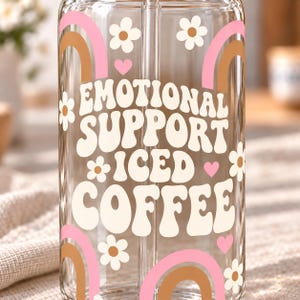 May include: A clear glass tumbler with a straw featuring the white text "EMOTIONAL SUPPORT ICED COFFEE" surrounded by pink and brown rainbow arches, small white flowers with brown centers, and tiny pink hearts printed on the exterior surface.