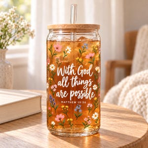 May include: A clear glass can-shaped tumbler with a bamboo lid and a glass straw, filled with amber liquid. The exterior features a colorful floral pattern and the white script text "With God all things are possible" and "MATTHEW 19:26".