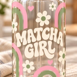 May include: A clear glass tumbler featuring the text MATCHA GIRL in large cream-colored letters. The design includes pink and sage green rainbow arches, small pink hearts, and white flowers with sage green centers printed on the exterior surface.
