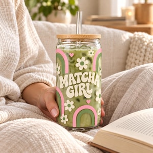 May include: A clear glass tumbler filled with a green matcha beverage, featuring a bamboo lid and a glass straw. The exterior is decorated with white flowers, pink hearts, and green and pink rainbows, with the text "MATCHA GIRL" in white.
