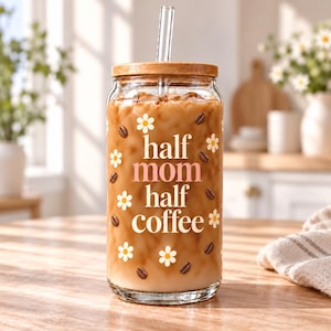 May include: A clear glass tumbler with a bamboo lid and a glass straw, filled with iced coffee. The exterior features white daisy icons, brown coffee bean graphics, and the text "half mom half coffee" printed in white and soft pink lettering.