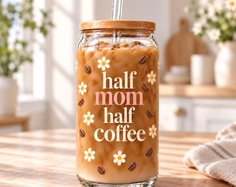 May include: A clear glass tumbler with a bamboo lid and a glass straw, filled with iced coffee. The exterior features white daisy icons, brown coffee bean graphics, and the text "half mom half coffee" printed in white and soft pink lettering.