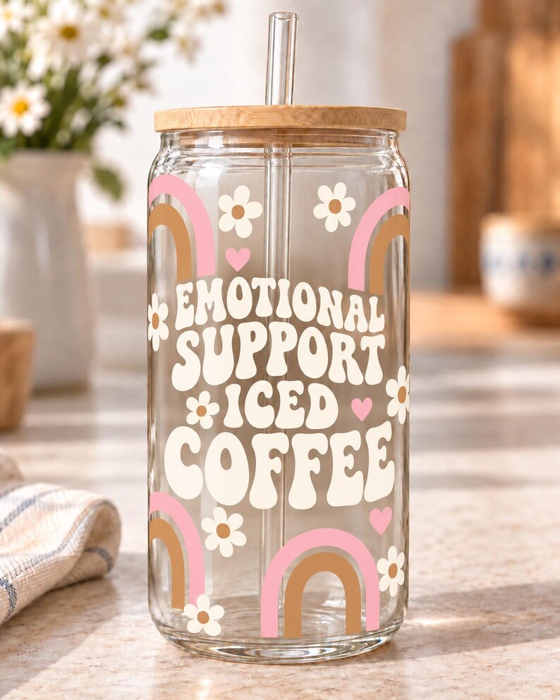 Digital mockup of a clear glass cup showing cream Emotional Support Iced Coffee lettering, pink and brown rainbows and white daisies. Bamboo lid shown.