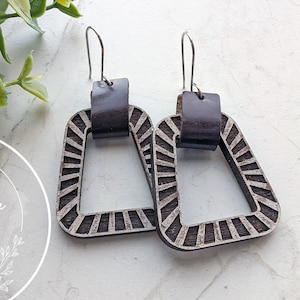 Könnte beinhalten: Ein Paar Holzohrringe mit geometrischem Design. Die Ohrringe sind braun und haben einen silbernen Haken. Die Ohrringe haben die Form eines Quadrats mit einem kleineren Quadrat, das aus der Mitte herausgeschnitten ist. Die Ohrringe hängen an einer weißen Oberfläche.