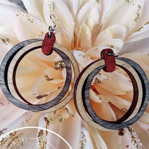 May include: Wooden hoop earrings with a layered design in shades of gray, brown, and natural wood. The earrings have a red accent piece at the top and are suspended from silver-colored ear wires. The earrings are displayed on a light pink flower.