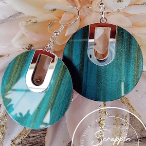 Op de afbeelding: Ronde houten oorbellen in teal en goud met een zilveren metalen accent. De oorbellen hebben een uniek ontwerp.