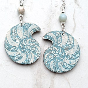 Peut inclure: Une paire de boucles d'oreilles avec un motif de coquille de nautile de couleur bleu et blanc. Les boucles d'oreilles sont en bois et ont des crochets et des perles argentés.