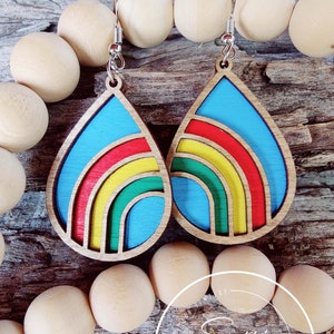 May include: A pair of teardrop-shaped wooden earrings with a rainbow design. The earrings are painted blue, red, yellow, and green. The earrings are hanging from silver hooks.