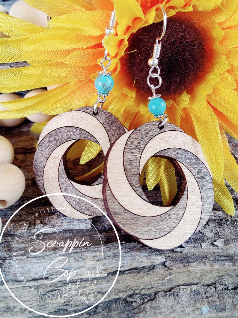 May include: A pair of wooden earrings with a spiral design. The earrings are made of light and dark wood and have a turquoise bead on each earring. The earrings are hanging from silver hooks.
