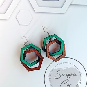 May include: A pair of wooden earrings with a layered hexagon design. The earrings are made of brown and green wood and have silver hooks. The earrings are shown against a white background with a circular logo that says "Scrappin' Cop" in a gray font.