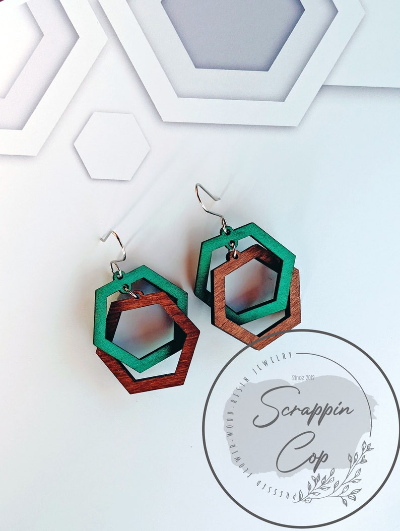 May include: A pair of wooden earrings with a layered hexagon design. The earrings are made of brown and green wood and have silver hooks. The earrings are shown against a white background with a circular logo that says "Scrappin' Cop" in a gray font.
