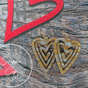Puede incluir: Un par de pendientes de coraz&oacute;n dorados con un dise&ntilde;o en capas. Los pendientes est&aacute;n hechos de un material transparente con detalles dorados. Los pendientes est&aacute;n colgados de una superficie de madera.