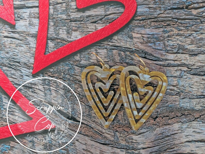 Puede incluir: Un par de pendientes de coraz&oacute;n dorados con un dise&ntilde;o en capas. Los pendientes est&aacute;n hechos de un material transparente con detalles dorados. Los pendientes est&aacute;n colgados de una superficie de madera.