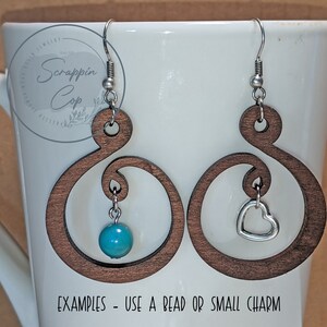 Pode incluir: Dois brincos de madeira com um design de espiral decorativo. Um brinco tem uma conta turquesa pendurada nele, e o outro tem um charme de cora&ccedil;&atilde;o prateado pendurado nele. "Exemplos - Use uma conta ou um pequeno charme" est&aacute; escrito abaixo dos brincos.
