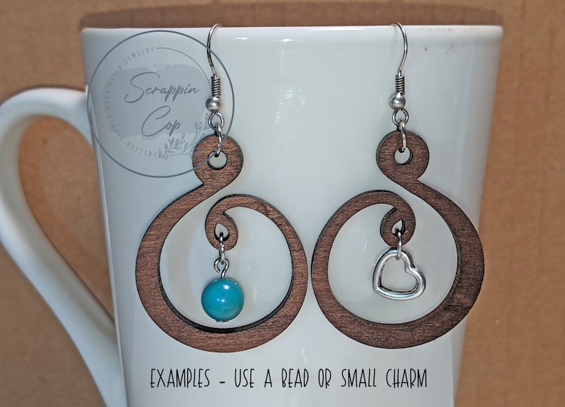 Pode incluir: Dois brincos de madeira com um design de espiral decorativo. Um brinco tem uma conta turquesa pendurada nele, e o outro tem um charme de cora&ccedil;&atilde;o prateado pendurado nele. "Exemplos - Use uma conta ou um pequeno charme" est&aacute; escrito abaixo dos brincos.