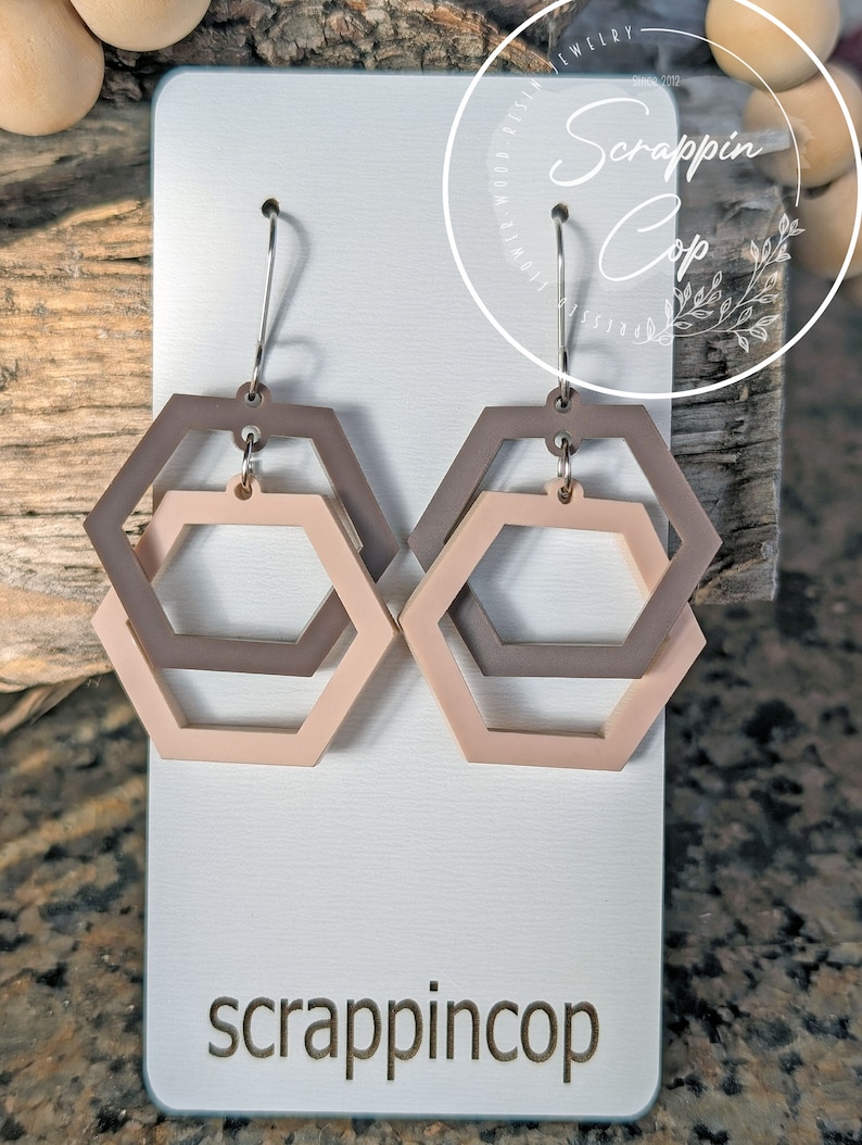 May include: A pair of earrings with a double hexagon design. The hexagons are in shades of brown and pink. The earrings are on a white card with the text "scrappincop" printed on it.