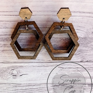 May include: A pair of wooden hexagon earrings with a layered design. The earrings are brown and have a natural wood finish. The earrings are hanging from a wooden post.