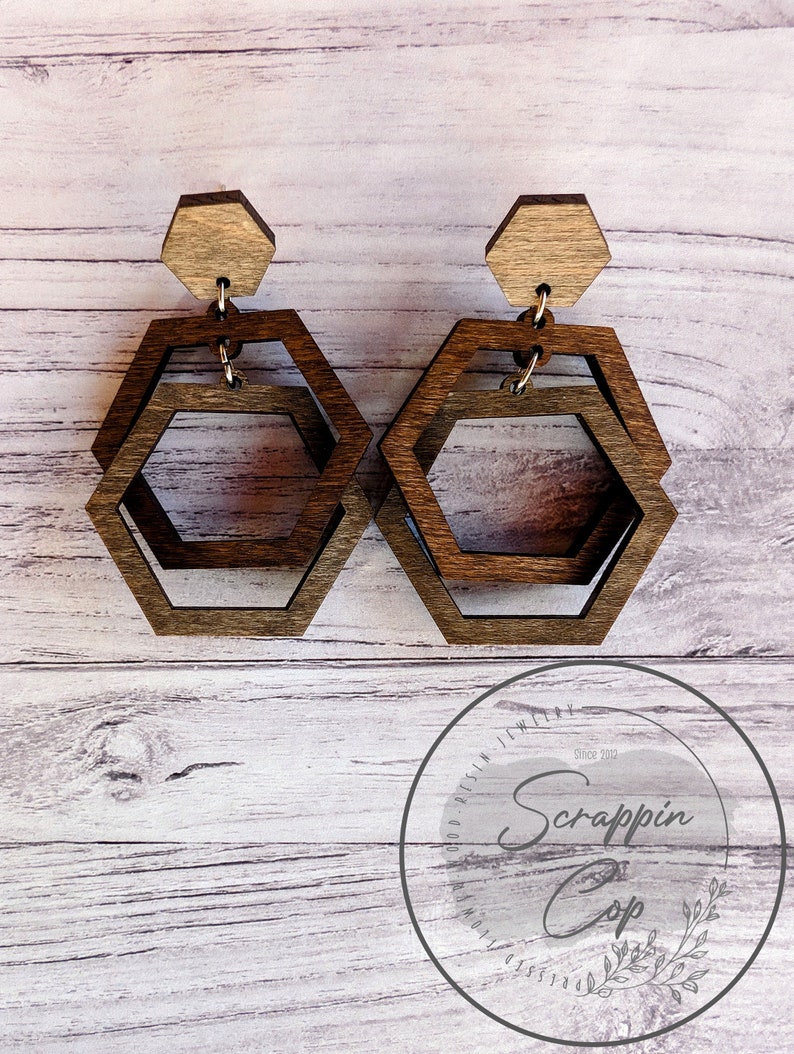 May include: A pair of wooden hexagon earrings with a layered design. The earrings are brown and have a natural wood finish. The earrings are hanging from a wooden post.