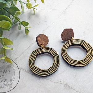 May include: A pair of wooden earrings with gold accents. The earrings are circular with a layered design and a small wooden hexagon attached to the top.