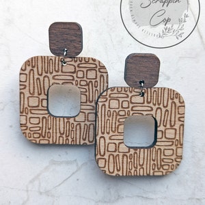 May include: A pair of wooden earrings with a geometric pattern. The earrings are square shaped with a smaller square cut out in the center. The earrings are made of light brown wood with a dark brown wood top.