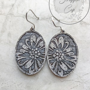 Könnte beinhalten: Ein Paar silberne Ohrringe mit einem weißen Blumenmuster. Die Ohrringe sind oval geformt und haben einen Used-Look.