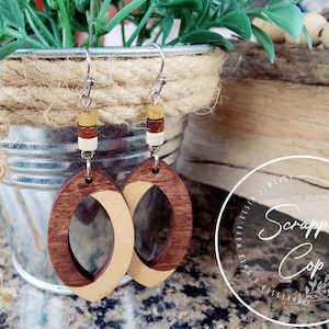 May include: A pair of wooden earrings with a light brown and dark brown wood design. The earrings are oval shaped and have a small hole in the center. The earrings are hanging from silver hooks with small wooden beads in brown, white, and yellow.