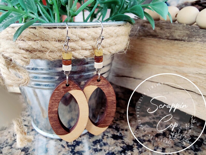 May include: A pair of wooden earrings with a light brown and dark brown wood design. The earrings are oval shaped and have a small hole in the center. The earrings are hanging from silver hooks with small wooden beads in brown, white, and yellow.