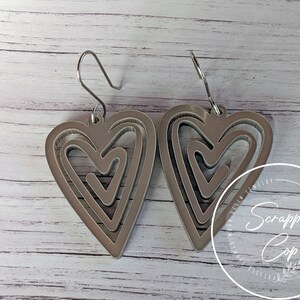 Puede incluir: Un par de pendientes de plata con un dise&ntilde;o de coraz&oacute;n en capas. Los corazones est&aacute;n superpuestos, creando un efecto tridimensional.