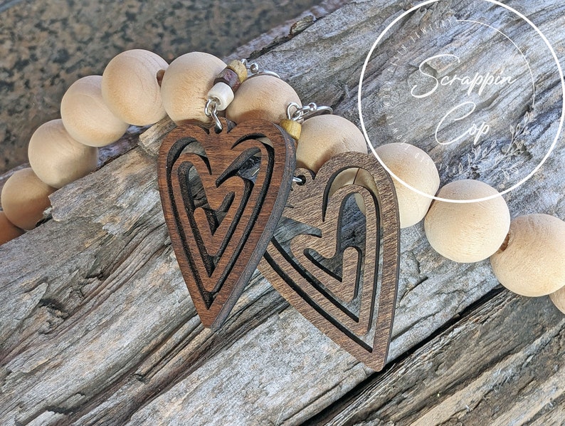 Puede incluir: Un par de pendientes de madera en forma de coraz&oacute;n con un dise&ntilde;o en capas. Los pendientes son marrones y tienen un acabado de madera natural. Est&aacute;n colgados de ganchos plateados.