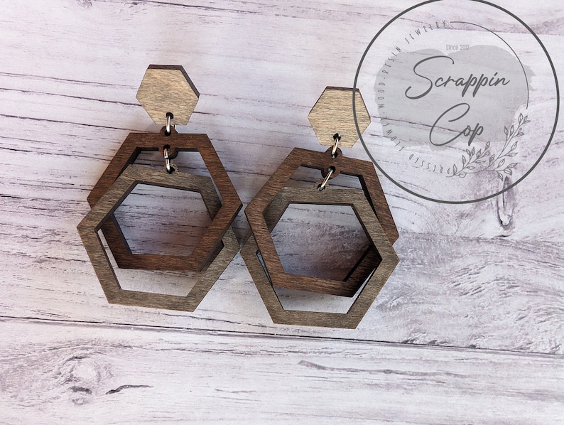May include: A pair of wooden earrings with a hexagonal shape. The earrings are made of two layers of wood, with the top layer being a lighter color than the bottom layer.