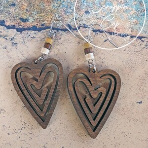 Puede incluir: Un par de pendientes de madera en forma de coraz&oacute;n con un dise&ntilde;o en capas. Los pendientes son marrones y tienen un acabado de madera natural. Est&aacute;n suspendidos de ganchos plateados con cuentas marrones y blancas.