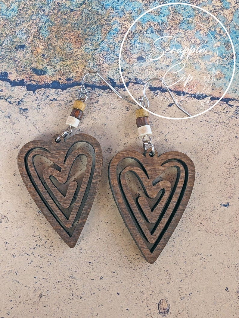Puede incluir: Un par de pendientes de madera en forma de coraz&oacute;n con un dise&ntilde;o en capas. Los pendientes son marrones y tienen un acabado de madera natural. Est&aacute;n suspendidos de ganchos plateados con cuentas marrones y blancas.