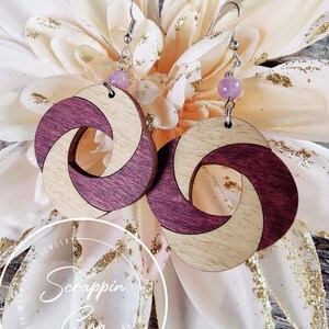 May include: A pair of wooden earrings with a swirl design. The earrings are made of light wood and dark wood, and they have a pink bead at the top of the earring hook.