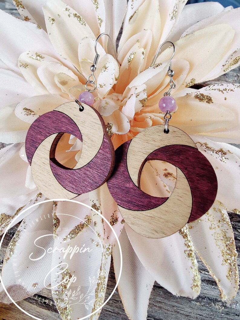 May include: A pair of wooden earrings with a swirl design. The earrings are made of light wood and dark wood, and they have a pink bead at the top of the earring hook.