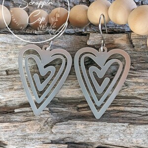Puede incluir: Un par de pendientes de plata en forma de coraz&oacute;n con un dise&ntilde;o abstracto en capas. Los pendientes est&aacute;n colgados de una superficie de madera.