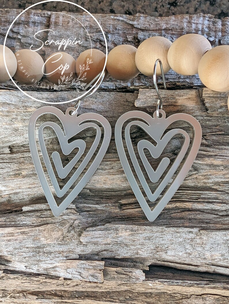 Puede incluir: Un par de pendientes de plata en forma de coraz&oacute;n con un dise&ntilde;o abstracto en capas. Los pendientes est&aacute;n colgados de una superficie de madera.