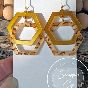 May include: Gold hexagon earrings with a leopard print design. The earrings are layered, with a smaller hexagon inside a larger hexagon.