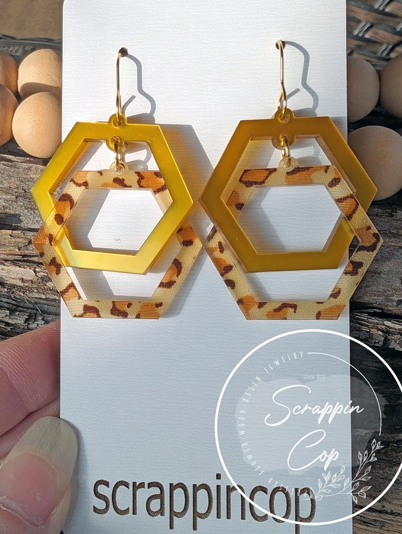 May include: Gold hexagon earrings with a leopard print design. The earrings are layered, with a smaller hexagon inside a larger hexagon.