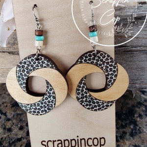 May include: A pair of wooden earrings with a leopard print design. The earrings are made of two pieces of wood that are twisted together. The earrings are hanging from silver hooks and have brown, white, and turquoise beads.