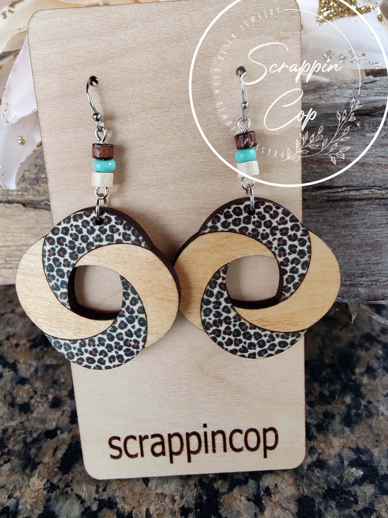 May include: A pair of wooden earrings with a leopard print design. The earrings are made of two pieces of wood that are twisted together. The earrings are hanging from silver hooks and have brown, white, and turquoise beads.