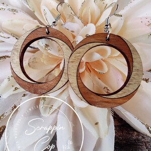 May include: A pair of wooden earrings with a natural wood finish. The earrings are circular and have a double-layered design. The earrings are hanging from silver hooks.