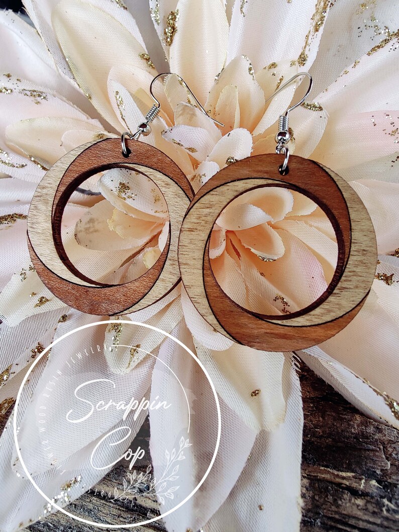 May include: A pair of wooden earrings with a natural wood finish. The earrings are circular and have a double-layered design. The earrings are hanging from silver hooks.